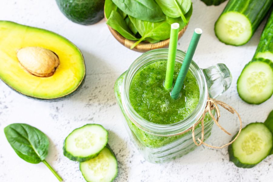 A green smoothie in a glass jar with straws next to avocado and vegetables